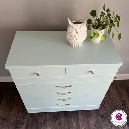 Light blue dresser with gold handles on a wooden floor, featuring decorative items including an owl-shaped vase and a potted plant.