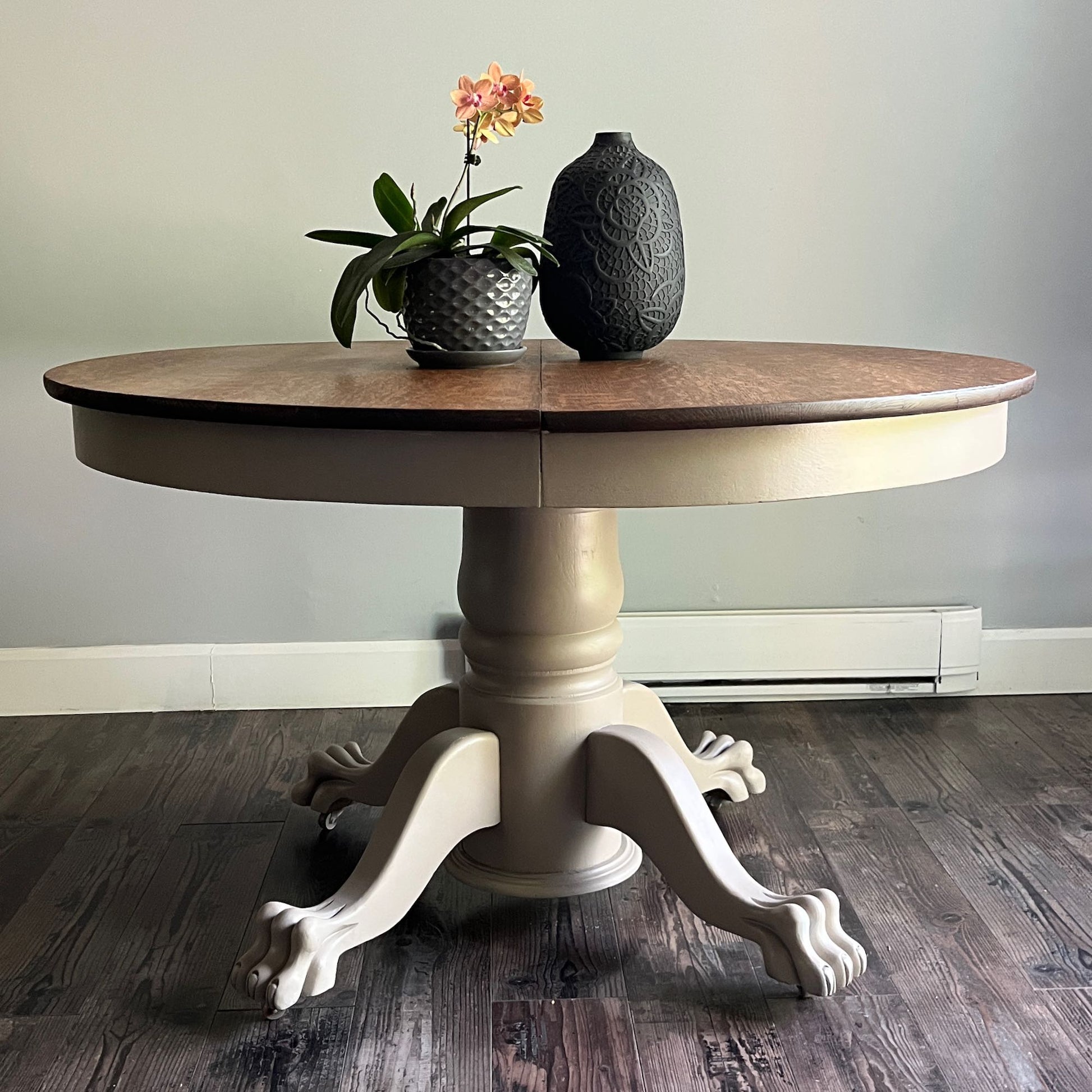 A round pedestal dining table with a medium brown wood top and a distressed beige/cream painted base with claw legs, shown on a dark wood floor against a light gray wall. Decorative items (a potted orchid and a textured vase) are placed on top.